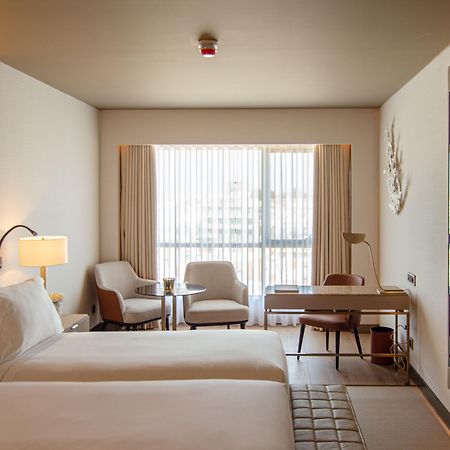 ליסבון Epic Sana Marques Hotel מראה חיצוני תמונה The photo depicts a modern hotel room featuring two beds with white linens. The room is well-lit, with large windows dressed in sheer curtains that allow natural light to filter in. There are two comfortable armchairs next to a small side table, prov