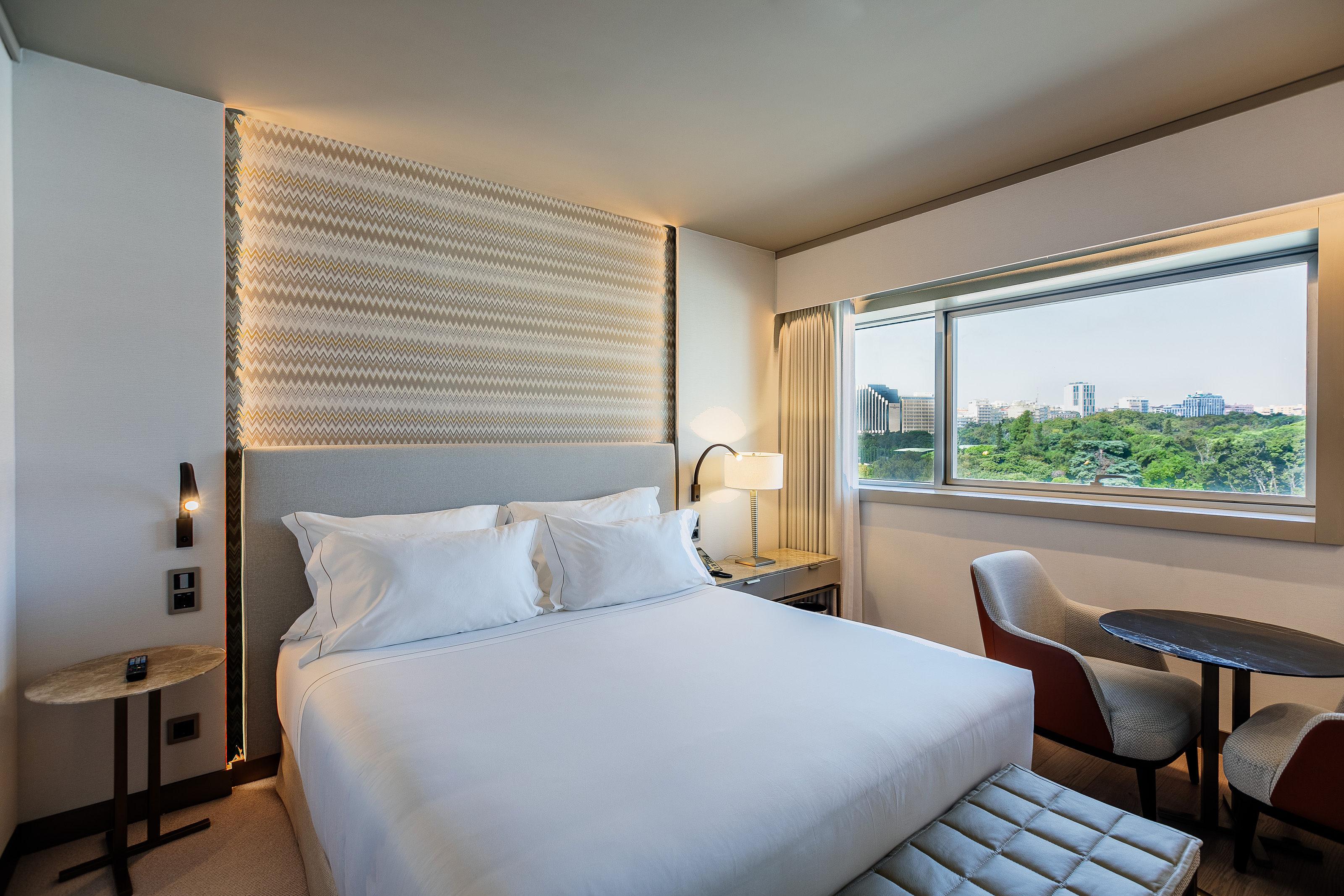 ליסבון Epic Sana Marques Hotel מראה חיצוני תמונה The photo shows a modern hotel room featuring a large bed with white linens and a padded headboard. There is a window that offers a view of greenery outside, suggesting a pleasant outdoor setting. Next to the bed, there is a bedside table with a lamp