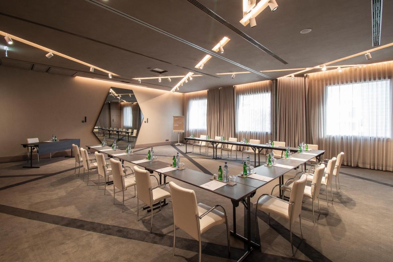 ליסבון Epic Sana Marques Hotel מראה חיצוני תמונה The photo shows a modern conference room with a sleek design. It features rectangular tables arranged in a U-shape, surrounded by white chairs. There are several bottles of water placed on the tables. The room has large windows with light curtains th